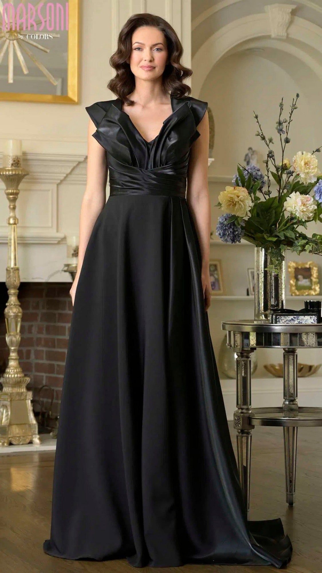 A woman in the Long Formal Dress: Marsoni M345, Black Black, stands indoors by a floral arrangement and fireplace, facing the camera.